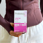 Une main d'une femme avec un sweatshirt couleur aubergine tient une boîte de Probiolog ventre plat devant son ventre