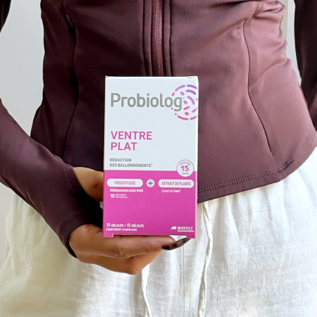 Une main d'une femme avec un sweatshirt couleur aubergine tient une boîte de Probiolog ventre plat devant son ventre