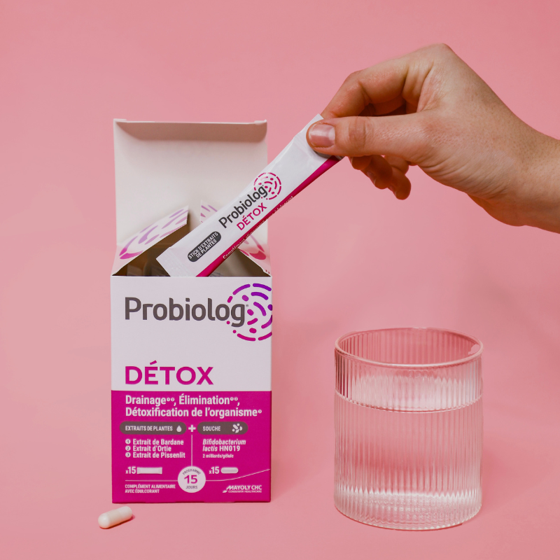 Une main prend un sachet de Probiolog Détox de sa boite avec un comprimé sur le côté et un grand verre d'eau