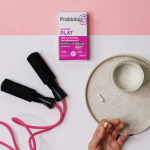 Boîte de Probiolog Ventre Plat à côté d'une corde à sauté rose, un verre et une capsule posés sur une assiette en gres et une main tenant une capsule Probiolog Ventre Plat